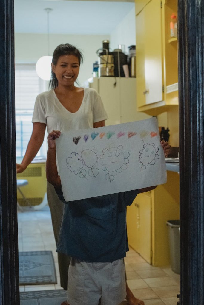 Child raising arms and showing drawing while standing near laughing young ethnic mother in light room